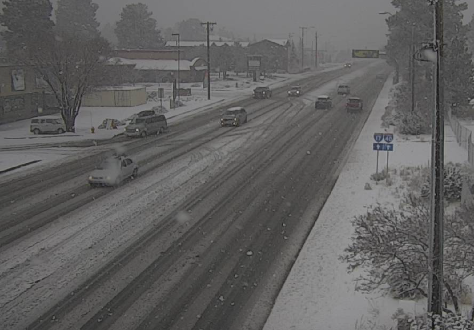Several cars drive on a snow-covered road during snowfall, with light snow accumulation visible on the ground, trees, and buildings.