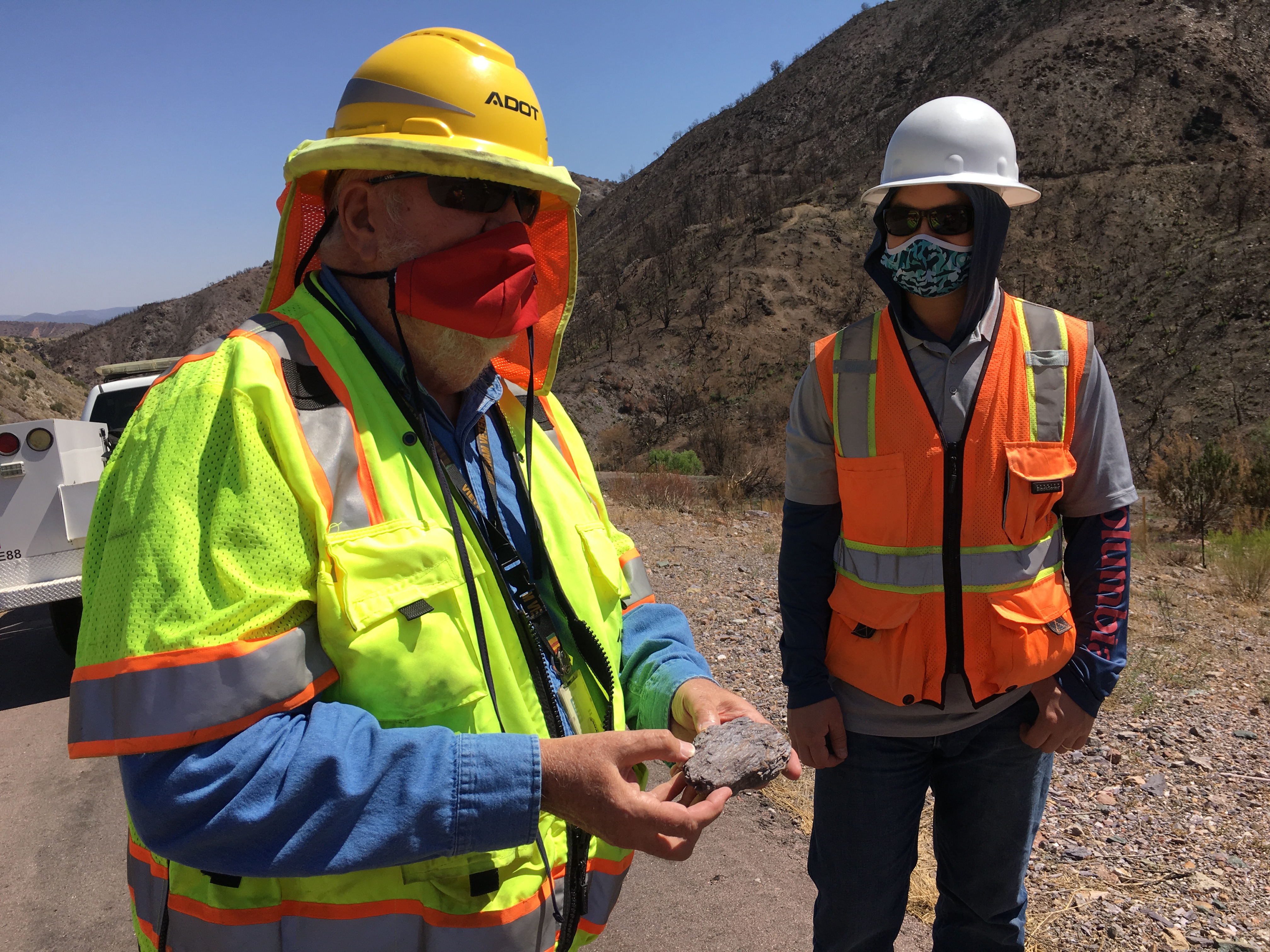 Two workers in safety vests and helmets, one holding a rock sample, stand on a roadside with rocky hills in the background. Both are wearing face masks.