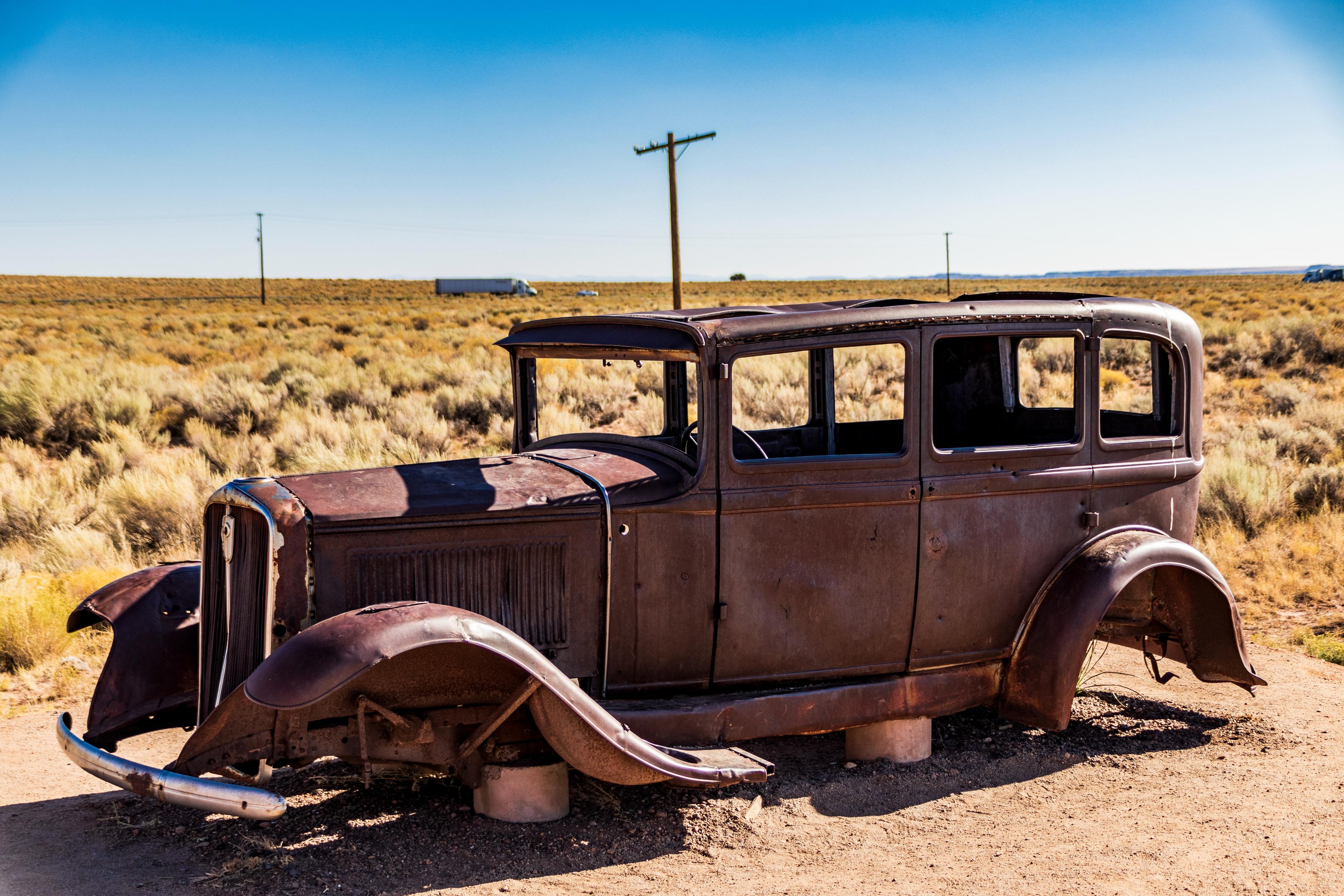 A rusted, vintage car sits abandoned in a dry, grassy landscape under a clear blue sky.