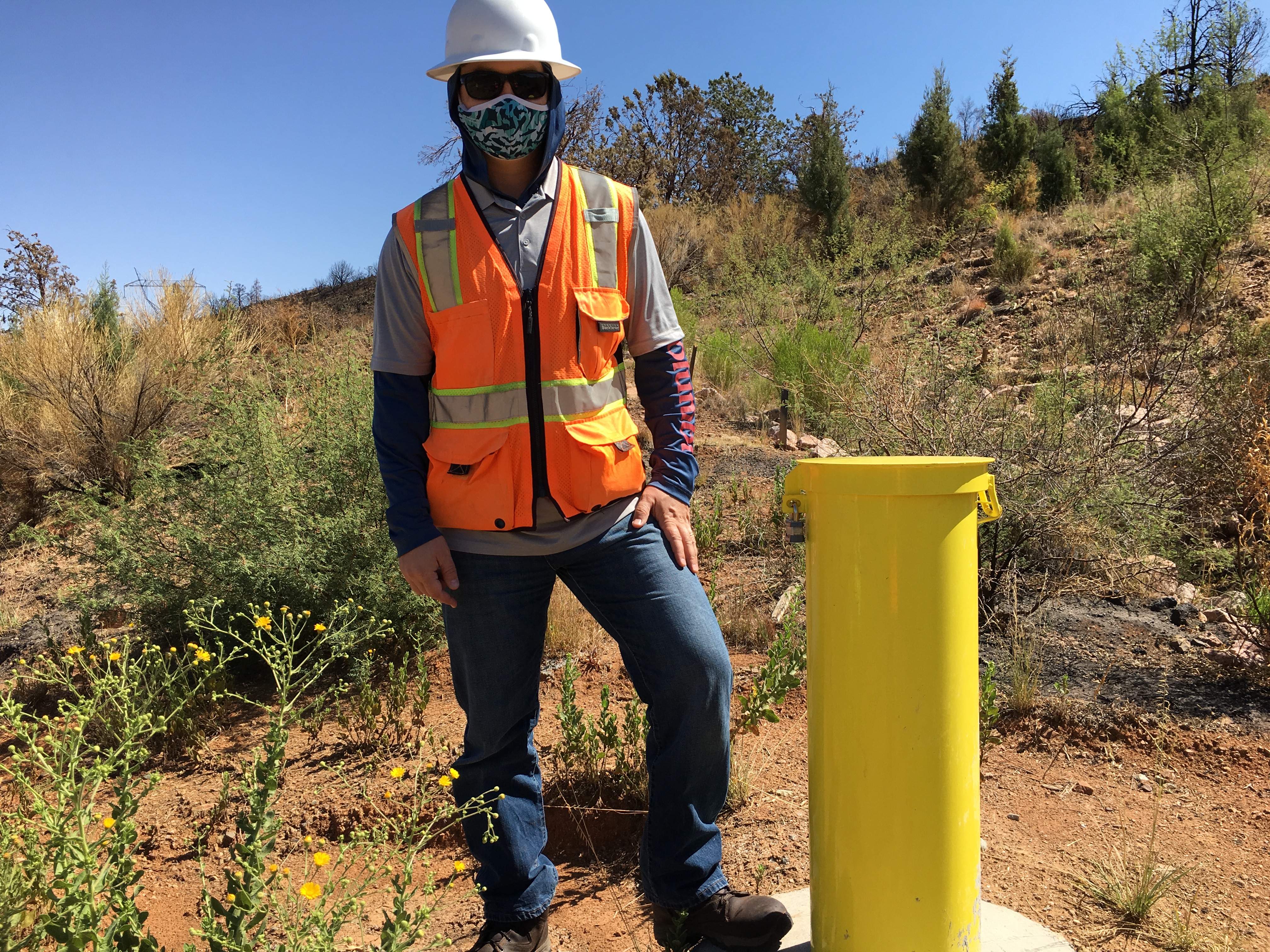 Person wearing a hard hat, safety vest, mask, and sunglasses stands outdoors next to a yellow post in a dry, brushy area.