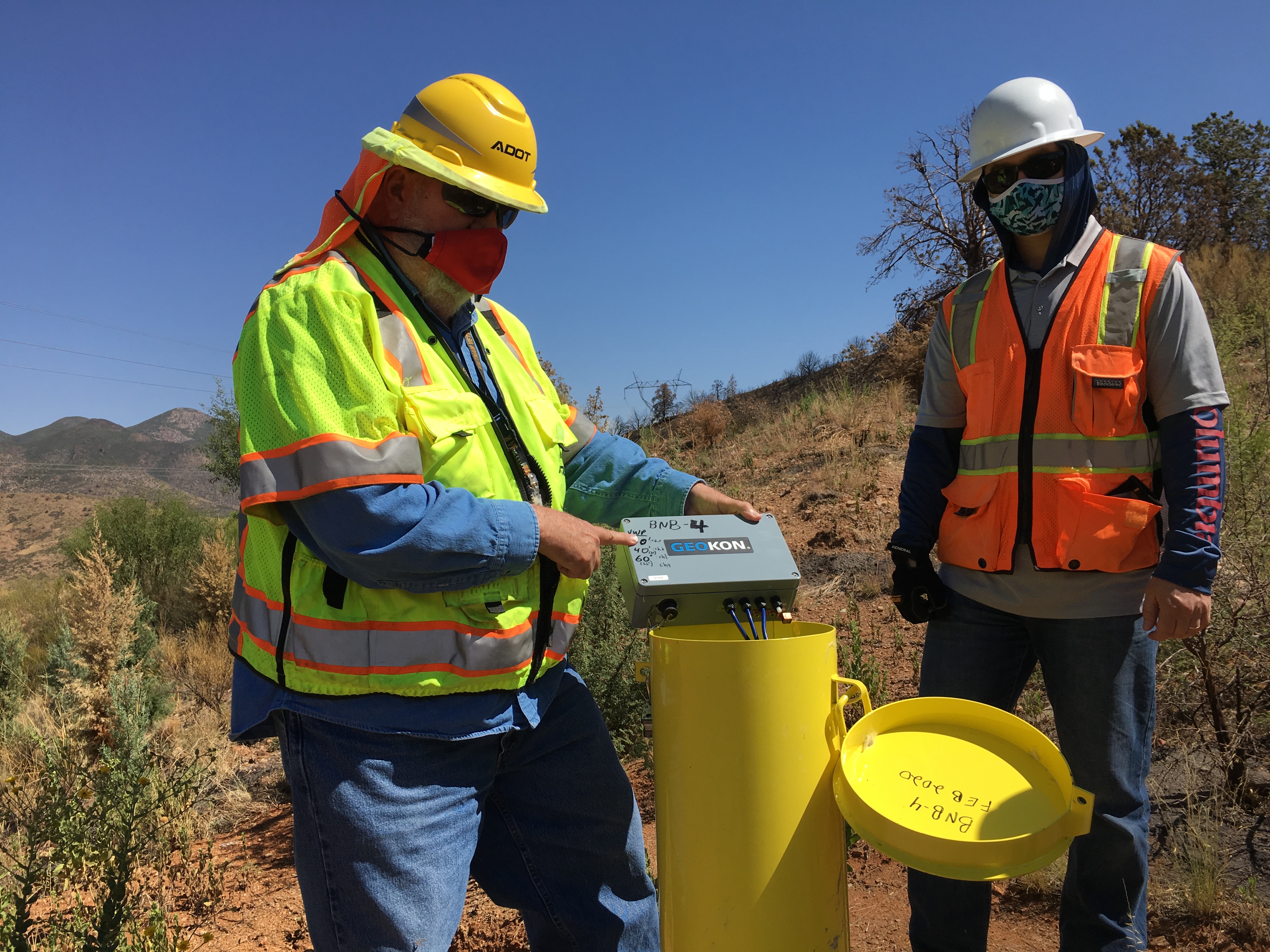 Two workers in safety vests and helmets operate a monitoring device at a yellow site marker in a dry, hilly outdoor area.