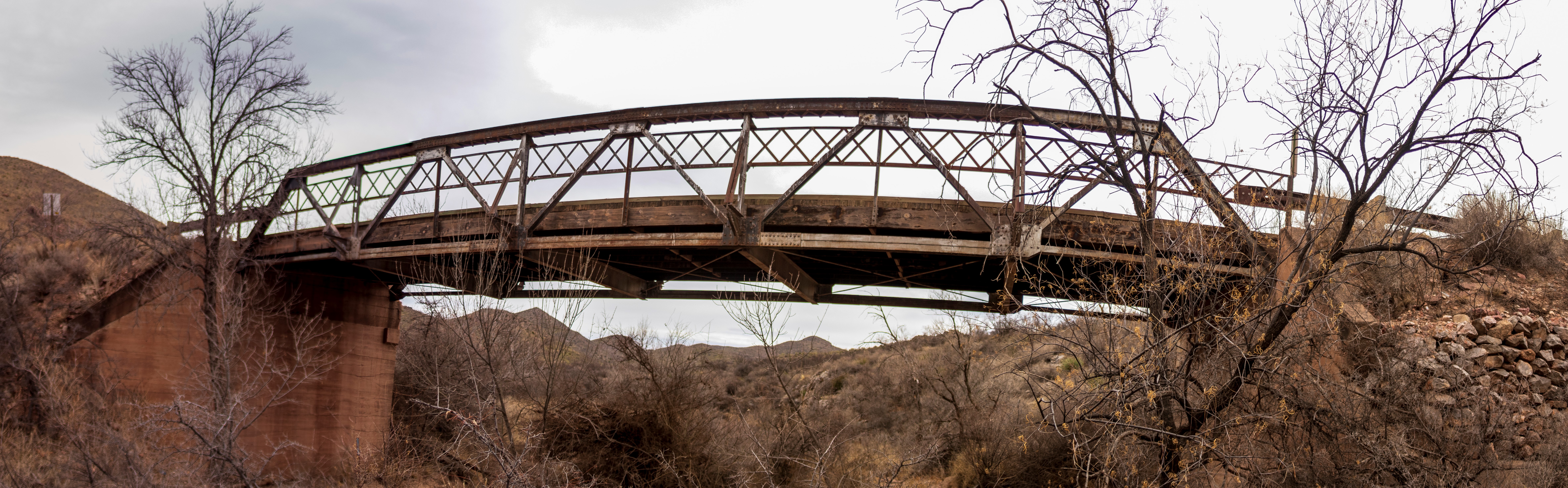 A weathered metal truss bridge the Leslie Creek Bridge spans a dry, brush-filled ravine with barren trees and distant hills under a cloudy sky.