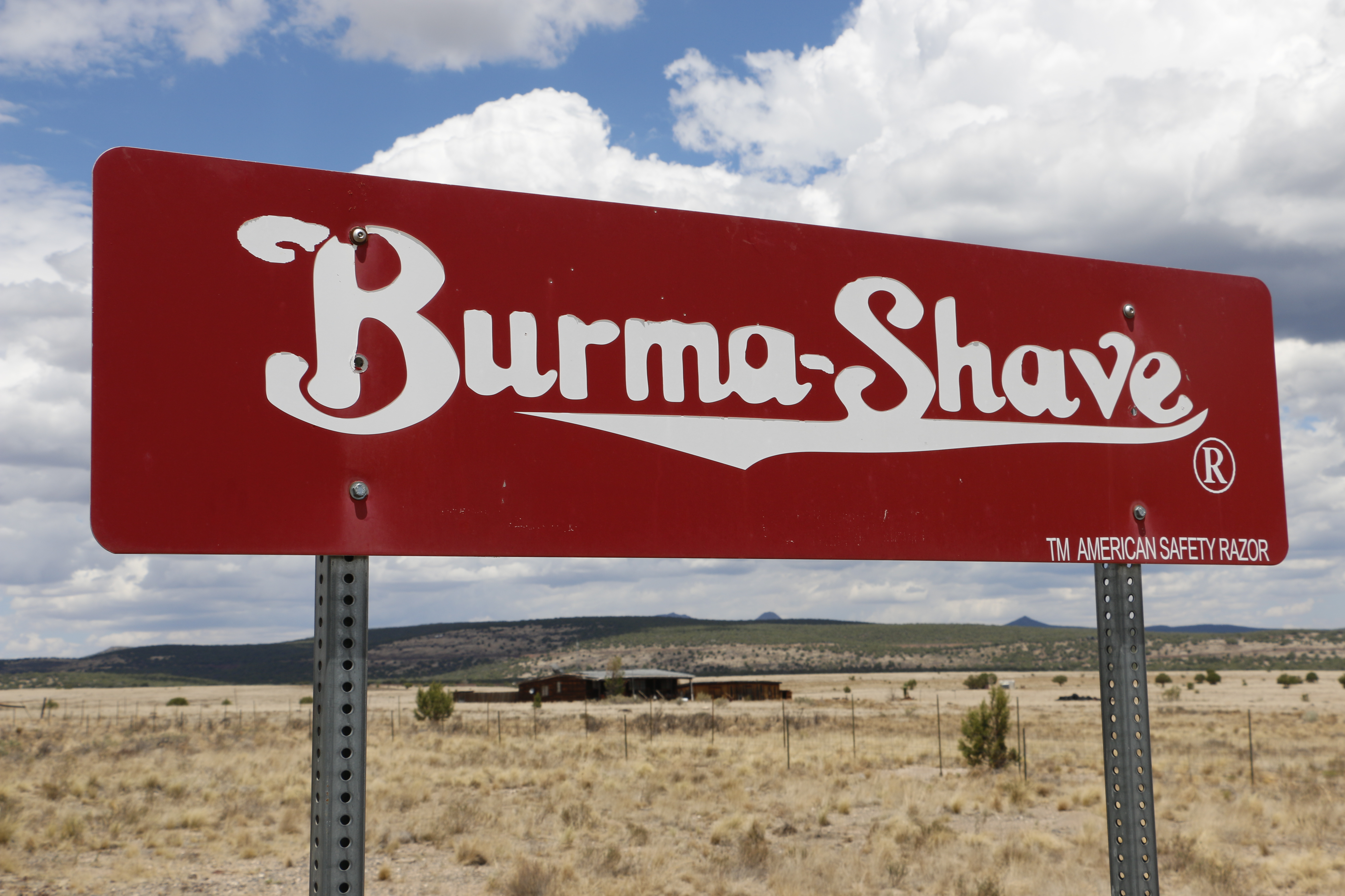 Red Burma-Shave sign stands in a dry, open landscape under a partly cloudy sky.