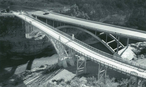 Two parallel bridges span a river, one modern with smooth arches, the other older with intricate truss design.