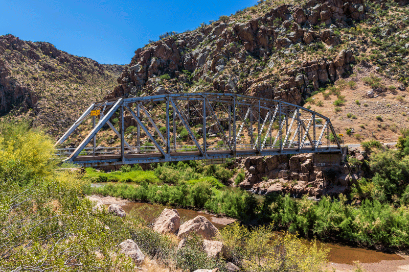 Steel truss bridge spans a rocky canyon with green shrubs and a river below, under a clear blue sky.