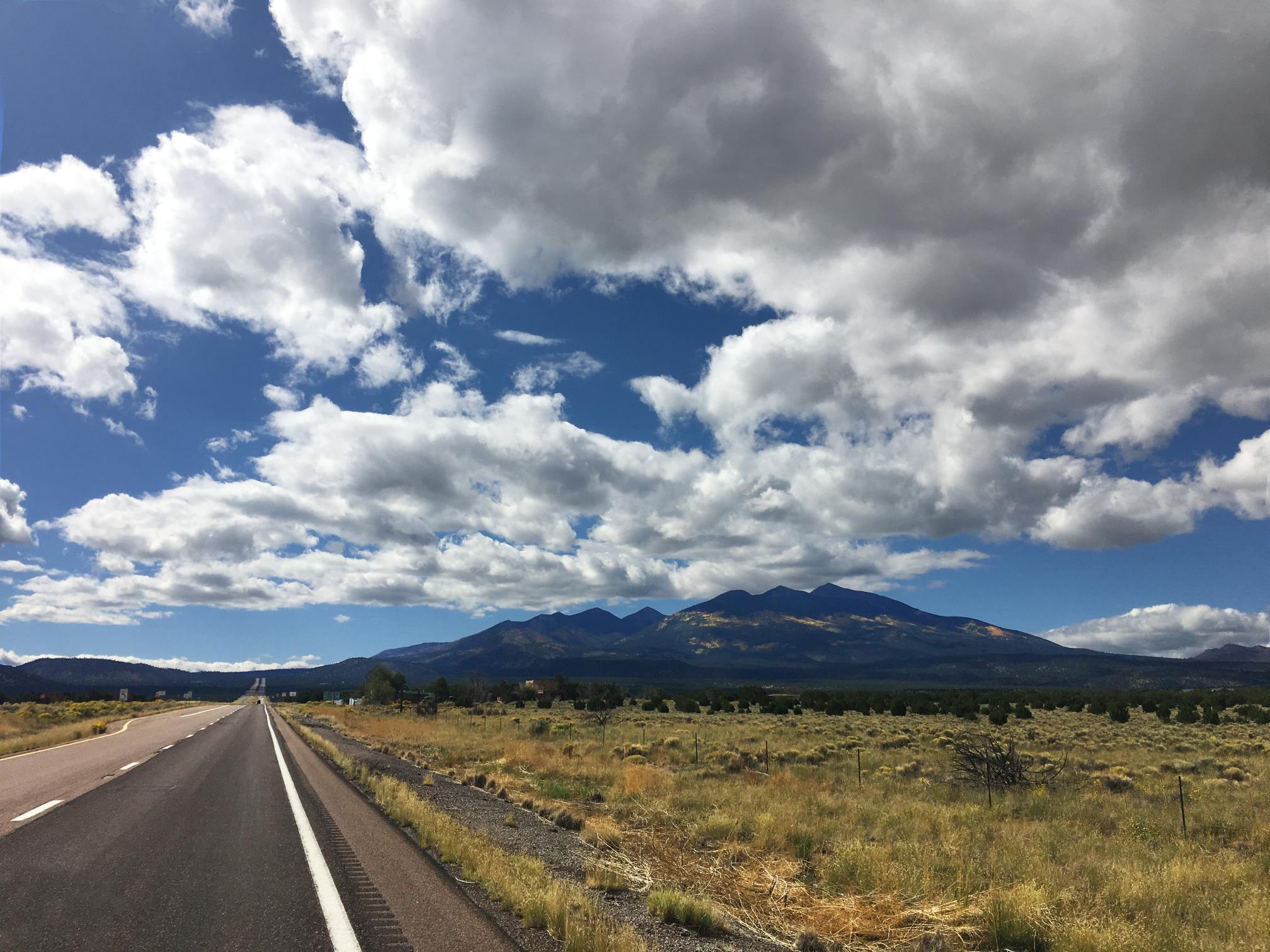 A highway runs toward distant mountains under a blue sky filled with large, fluffy clouds.