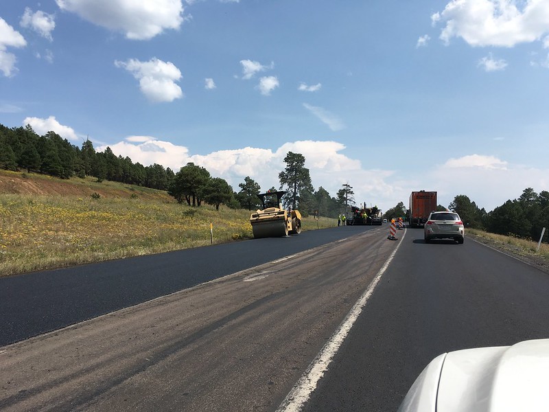 Road construction with vehicles and workers paving a rural highway under a partly cloudy sky.