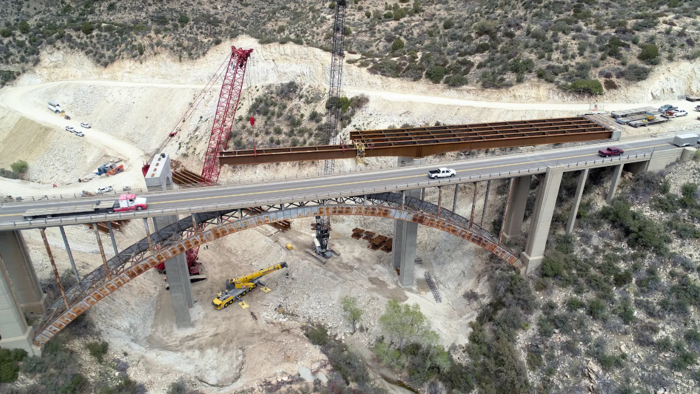 Aerial view of the Pinto Creek Bridge in Arizona under construction with cranes, trucks, and equipment in a hilly area.