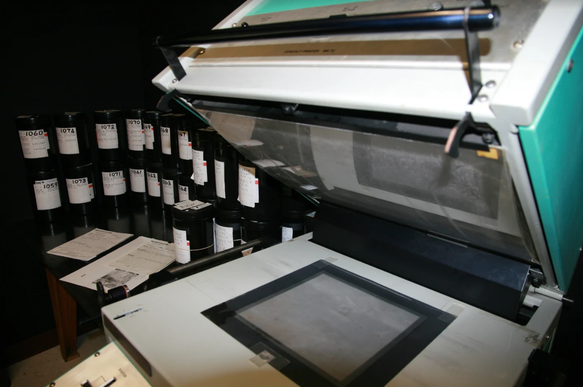 A microfilm reader with reels of microfilm and papers on a nearby table in a dimly lit room.