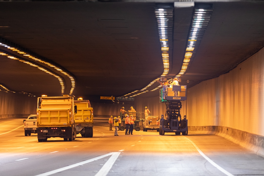 Road workers in safety vests repair lights in a tunnel with trucks and a lift blocking traffic lanes.