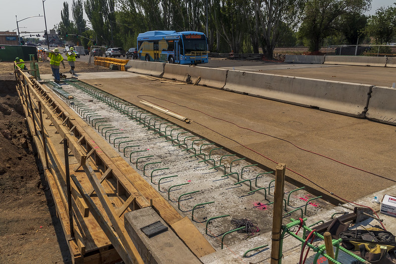 Construction workers build the Rio de Flag Bridge in Flagstaff Arizona. A public transit bus and traffic barriers are visible in the background.
