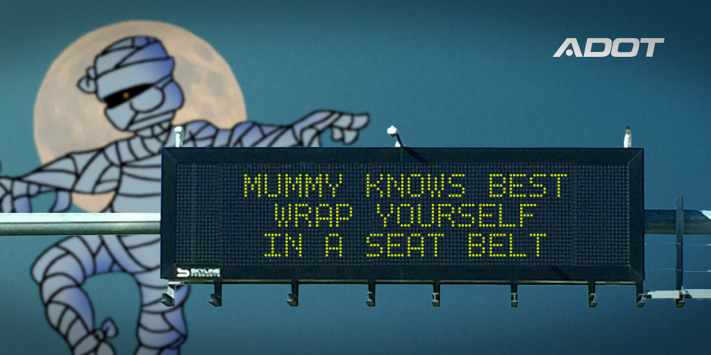 A highway sign reads Mummy knows best, wrap yourself in a seat belt with a cartoon mummy and a full moon in the background. ADOT logo appears in the top right corner.