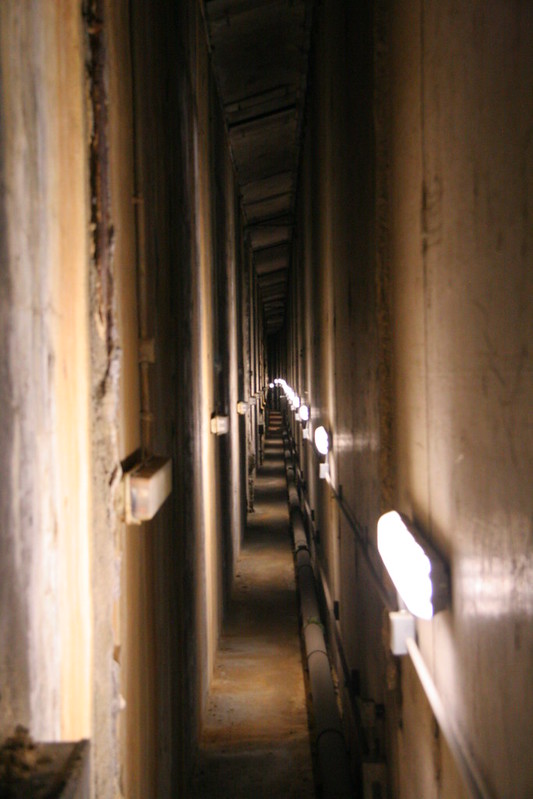 A narrow, dimly lit concrete corridor with exposed pipes and wall-mounted lights running along its length.