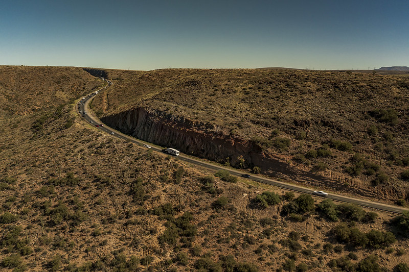 A winding road with cars cuts through a dry, hilly landscape under a clear blue sky.