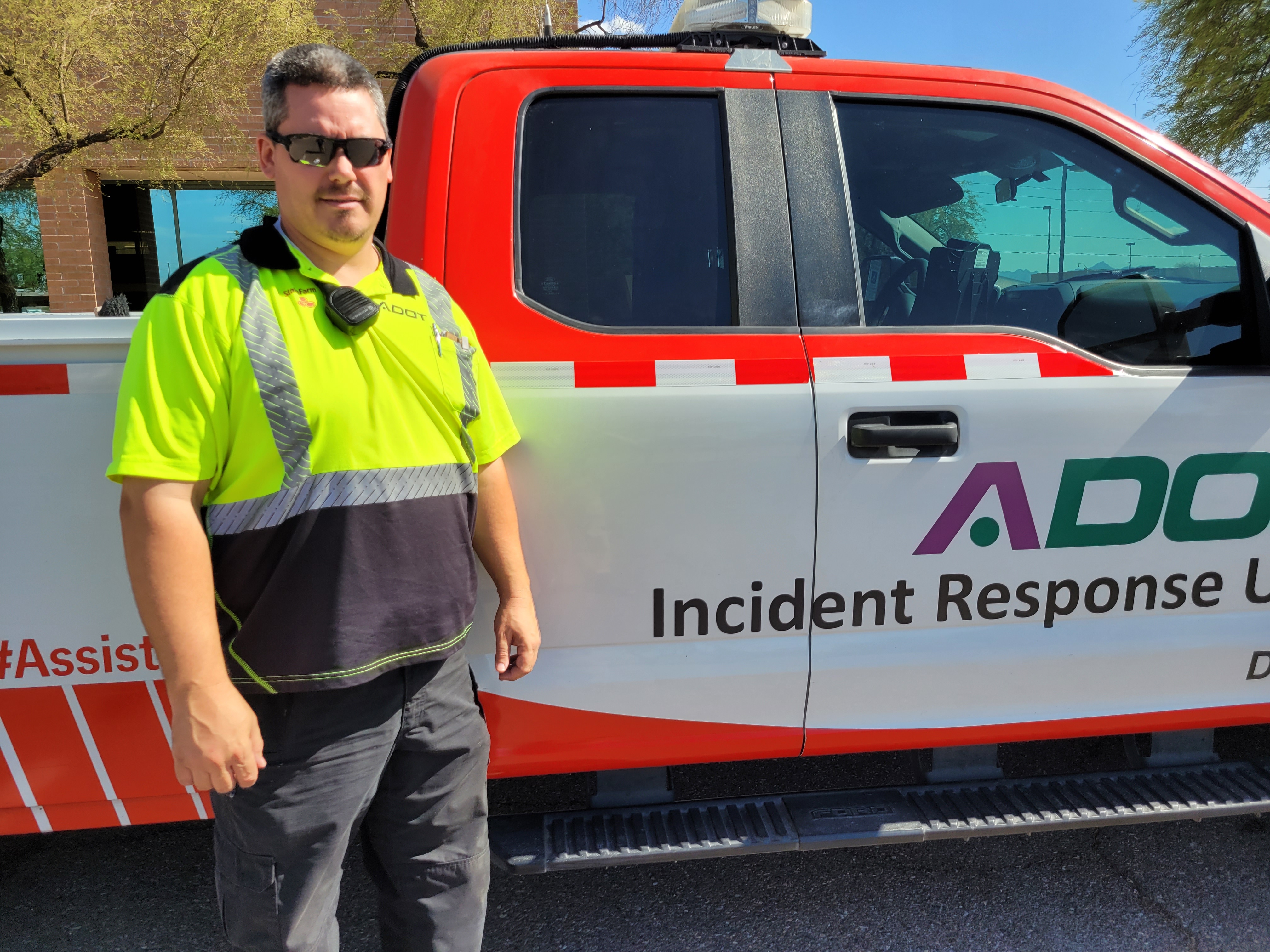 Man in a high-visibility shirt stands next to an Incident Response Unit truck in sunlight.