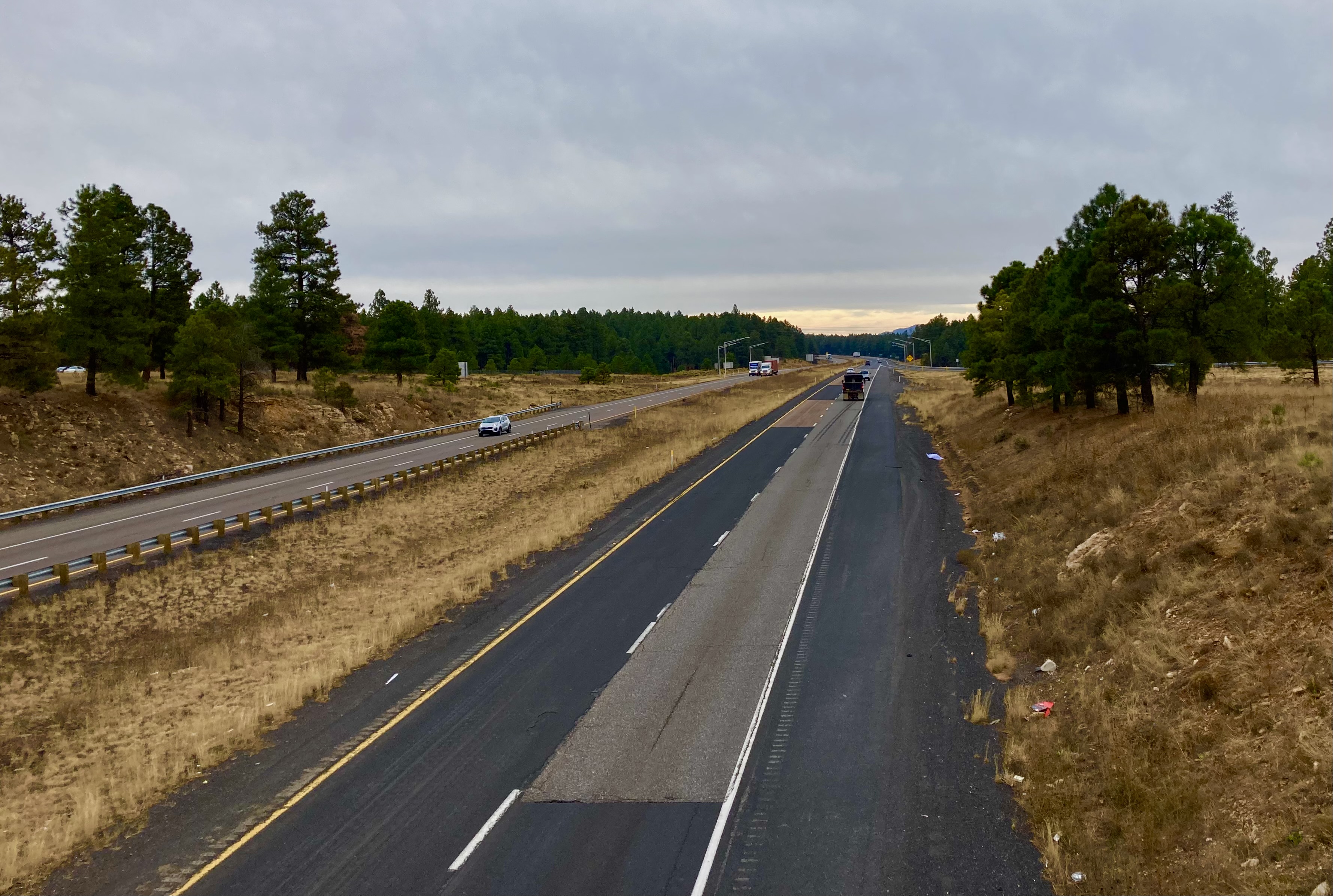 A divided highway with a few cars, bordered by trees and dry grass under a cloudy sky.