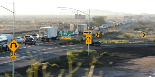 Cars and trucks are lined up in slow-moving traffic on a highway near an exit, with road signs indicating a 25 mph speed limit and a U-turn.