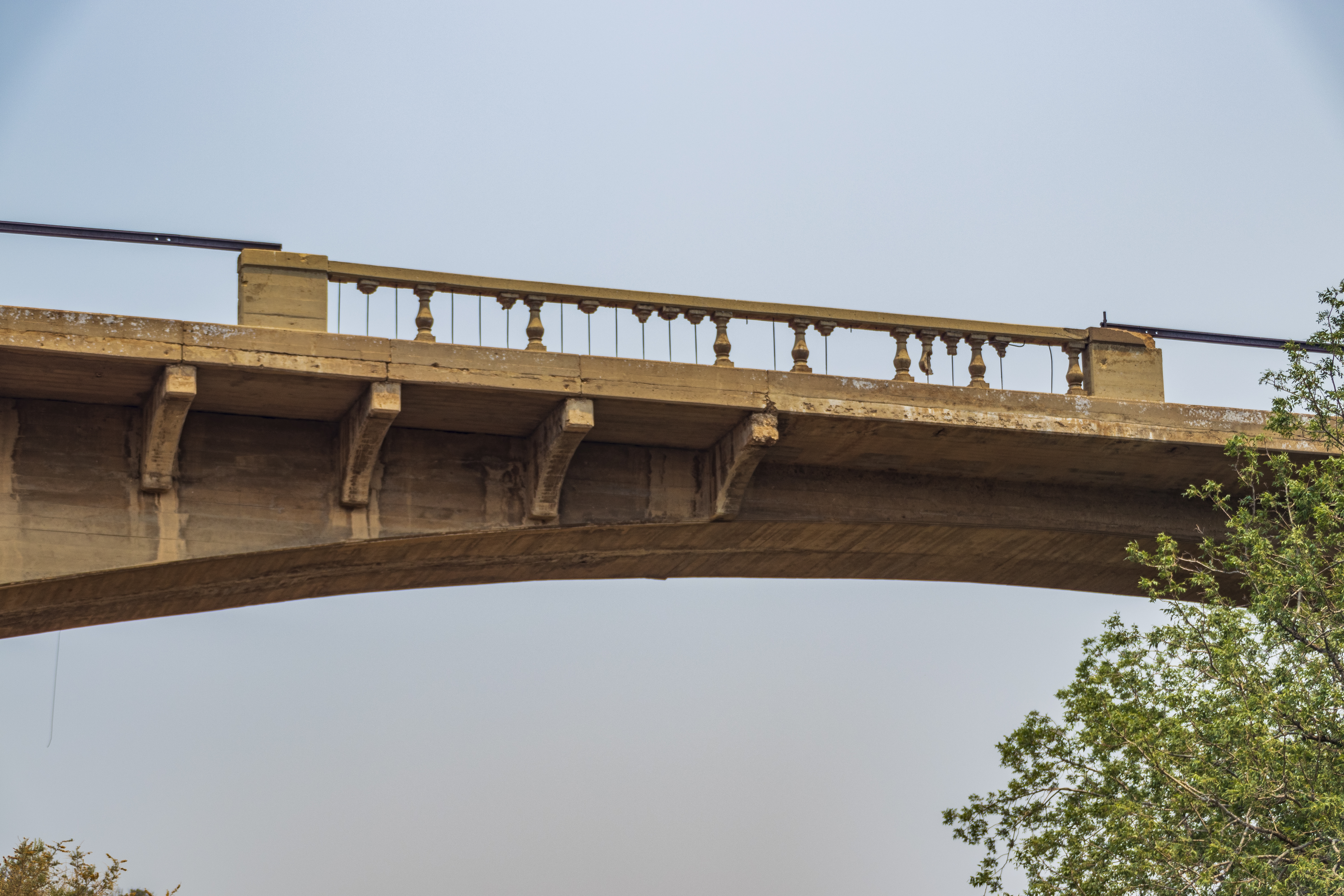 An old concrete bridge with visible railings and support beams, photographed from below against a clear sky with tree branches in the foreground.