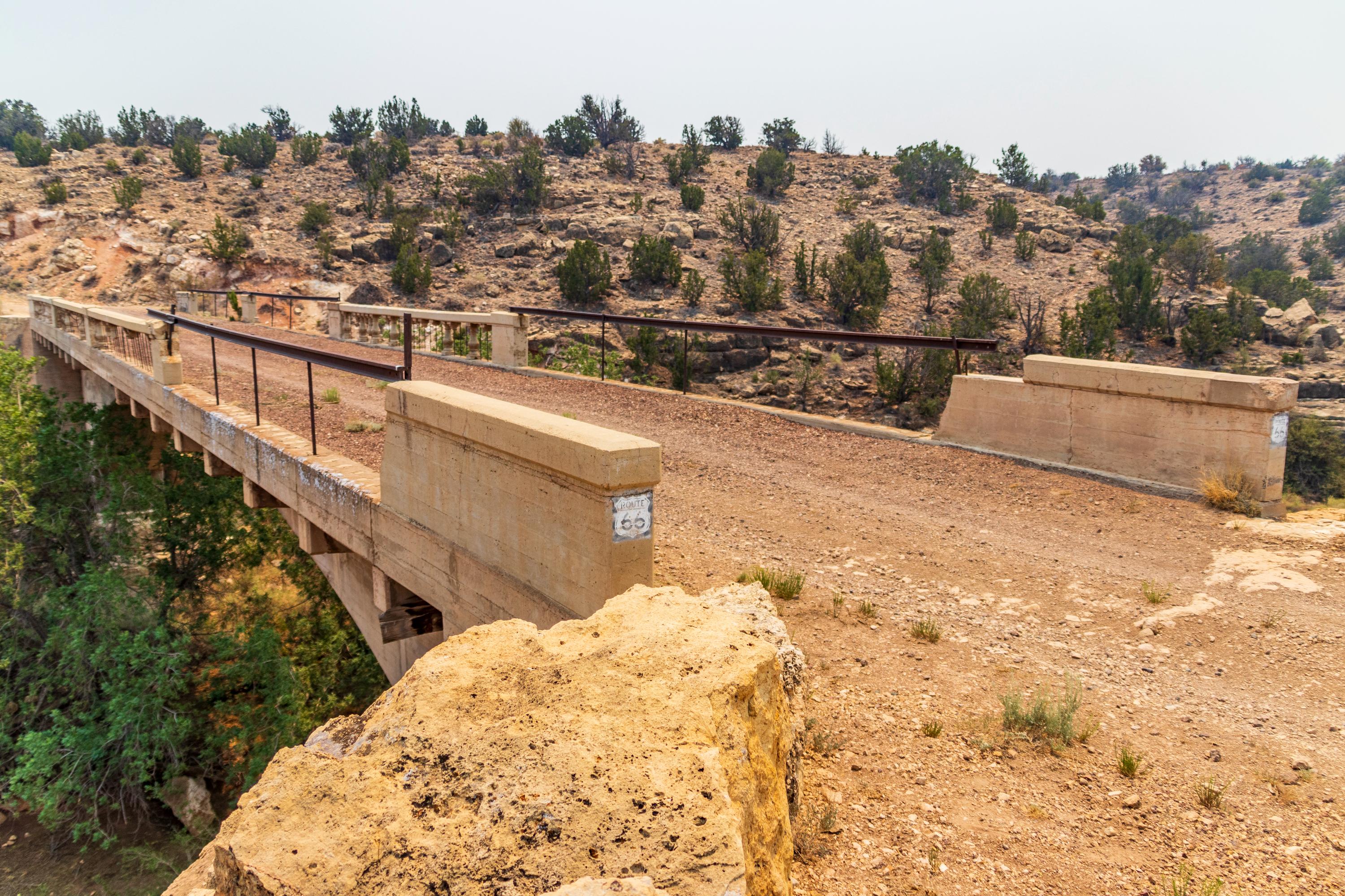 A narrow, weathered concrete bridge with metal railings crosses a dry, rocky landscape with sparse trees and shrubs.