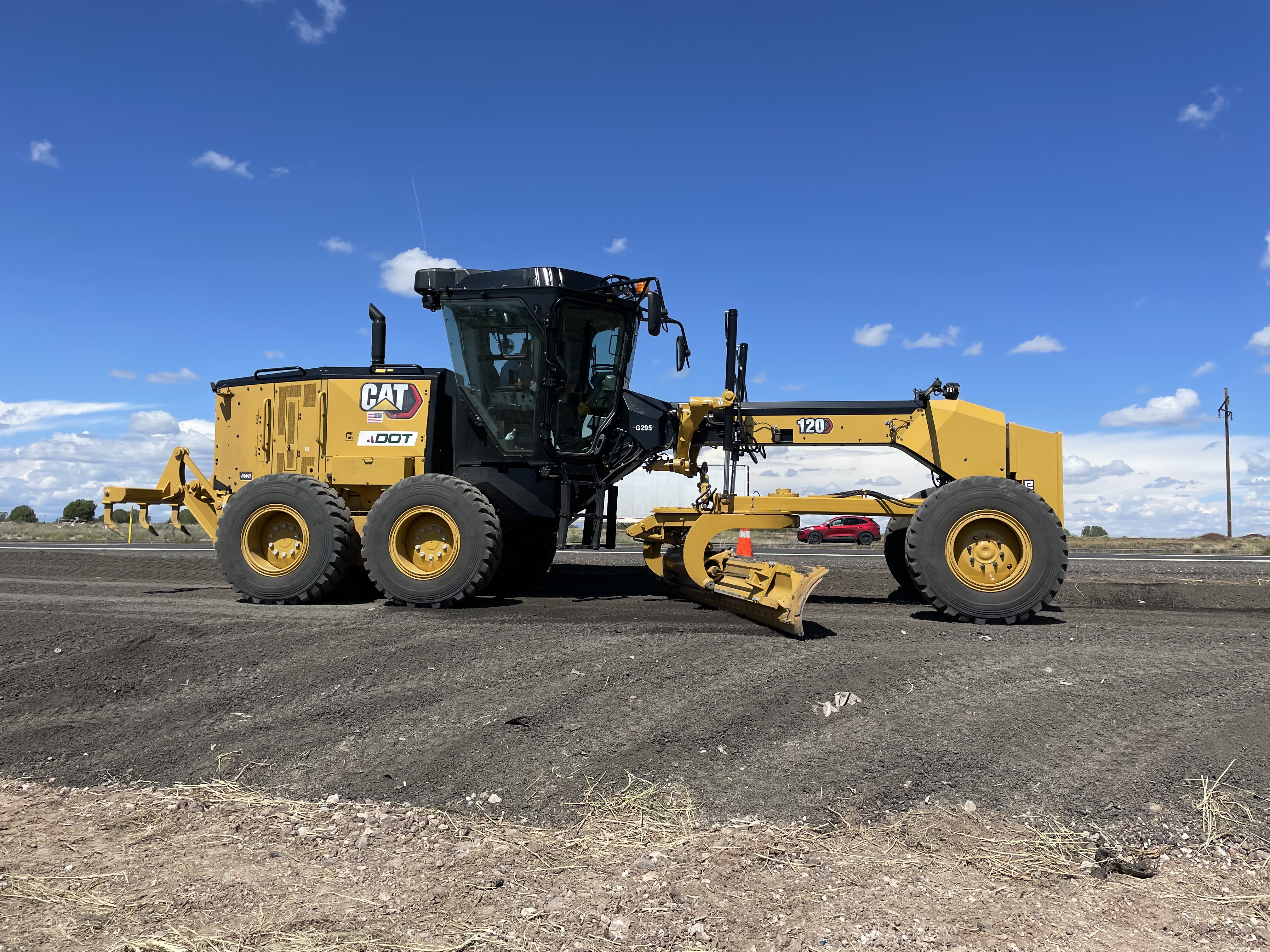 A yellow Caterpillar road grader working on a dirt road under a blue sky with scattered clouds.