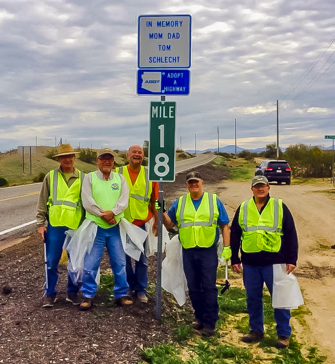 Five people in safety vests stand by a roadside Adopt A Highway sign dedicated to Tom Schlecht, holding trash bags near a mile marker on a cloudy day.