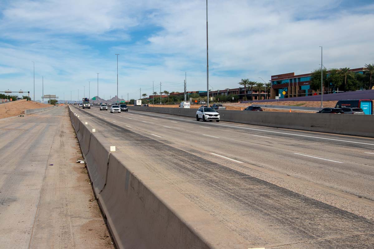 Traffic flowing on Loop 101 alongside concrete construction barriers and nearby commercial buildings.
