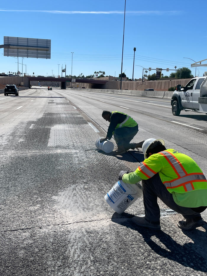 Construction workers applying crack-seal material to pavement on Loop 101, kneeling with buckets along freshly milled freeway lanes.