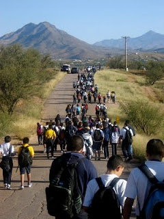 A large group of schoolchildren with backpacks walk down a rural road toward distant mountains.