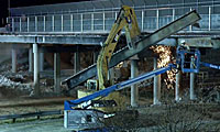 A construction worker uses a lift to cut a large metal beam with sparks flying, while an excavator supports the beam near a bridge at night.
