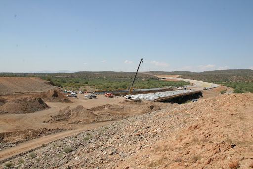 A construction site with vehicles, dirt piles, and a crane building a bridge over a road in a dry, hilly landscape under a clear sky.