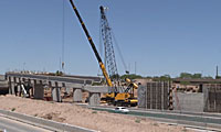 A crane lifts a large concrete section during bridge construction over a highway, with several support columns and a car driving below.