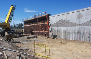 A construction site with a crane, scaffolding, caution tape, dirt ground, and a large unfinished concrete wall under a clear blue sky.