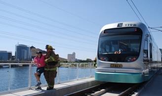 A firefighter assists a person off a metro light rail train near a waterfront on a sunny day.