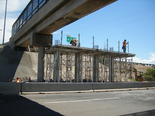 Two construction workers stand on scaffolding under an overpass, working on a concrete structure above a road with vehicles and equipment nearby.