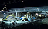 Construction equipment and vehicles work under a highway overpass at night, illuminated by overhead lights.