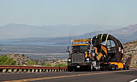 A large black semi-truck marked Oversize Load transports heavy equipment on a flatbed trailer along a highway with mountains in the distance.