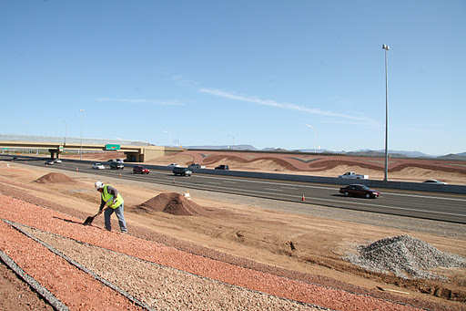 A construction worker in a yellow vest works on a dirt embankment beside a busy multi-lane highway under a clear blue sky.