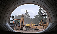 View from inside a large pipe showing construction vehicles, including a backhoe and bulldozer, working at a construction site with trees in the background.