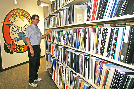 Man browsing binders on a library shelf near a wall with a Roadrunner Café cartoon mural.