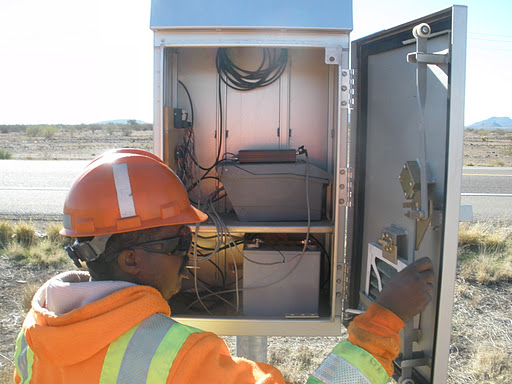 A person wearing an orange safety helmet and reflective jacket inspects the open control panel of a roadside electronic equipment box.