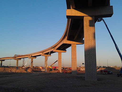 A large concrete highway overpass under construction, supported by tall pillars, with cranes and construction equipment on the dirt ground below.
