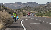 A person in a yellow vest walks along the shoulder of a highway, carrying a large blue bag, with cars and trucks on the road and mountains in the background.