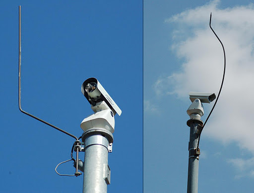 Two surveillance cameras are mounted on metal poles. The left cameras antenna is straight, while the right cameras antenna is bent upwards and twisted.