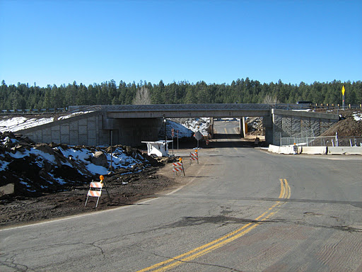 A concrete overpass bridge spans a two-lane road, with snow patches on the ground and construction signs placed along the roadside. Trees line the background.