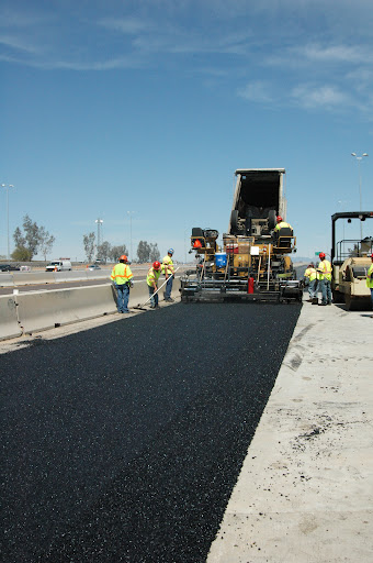Construction workers pave a highway using large machinery. Fresh asphalt is being laid, and workers in safety vests are standing on both sides of the road.