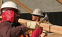 Two construction workers wearing hard hats and gloves hammer wooden beams at a building site.