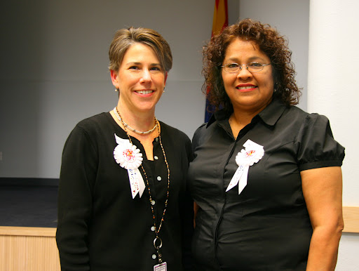 Two women stand side by side indoors, both wearing black tops and white ribbon badges, smiling at the camera.