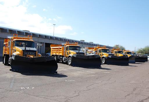 A row of yellow snow plow trucks are parked in a lot under a clear blue sky.