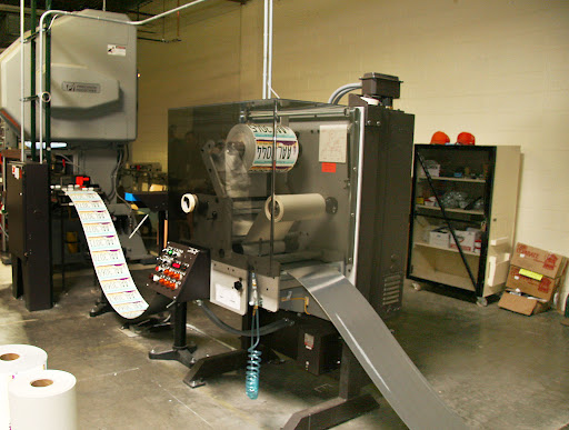 A license plate printing machine is producing a continuous roll of printed license plates from a sheet of steel. Shelves and boxes are visible in the background.