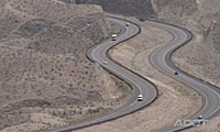A winding highway curves through a dry, hilly desert landscape with several vehicles traveling in both directions.